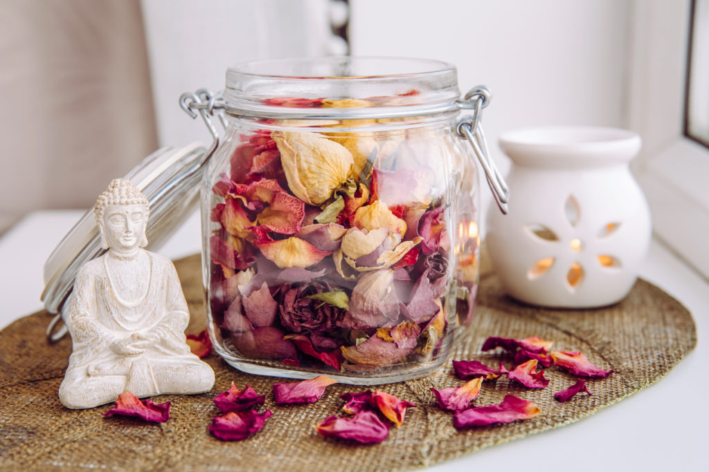 Mason jar filled with dry rose petals and buds with meditating Buddha figurine and aroma lamp with candle next to it.