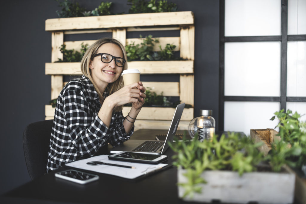 pretty, young blond businesswoman with a black and white checked shirt sits in a sustainable, ecological office and drinks coffee to go