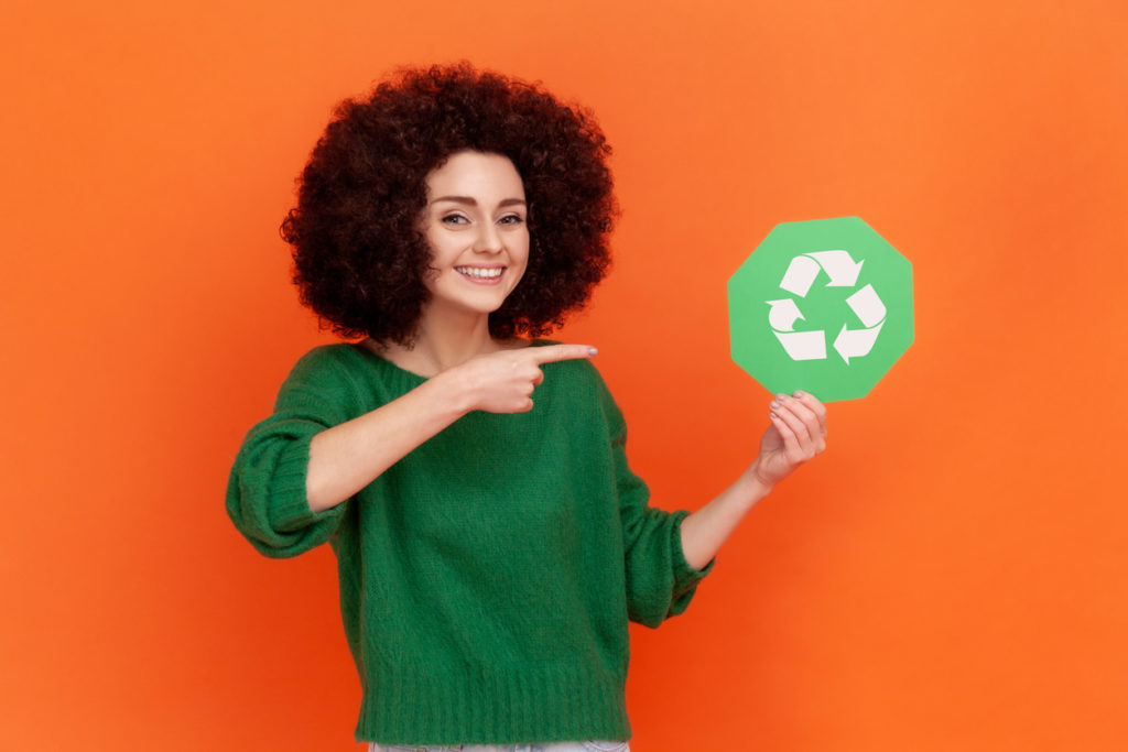 Satisfied woman with Afro hairstyle wearing green casual style sweater pointing at green recycling symbol in her hands, ecology concept. Indoor studio shot isolated on orange background.