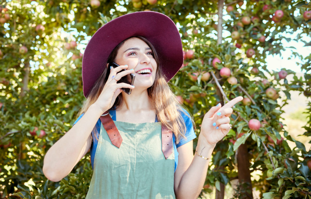 One happy woman talking on cellphone while working on sustainable apple orchard farm on sunny day. Cheerful farmer speaking on phone while planning tasks for harvest season of fresh organic fruit