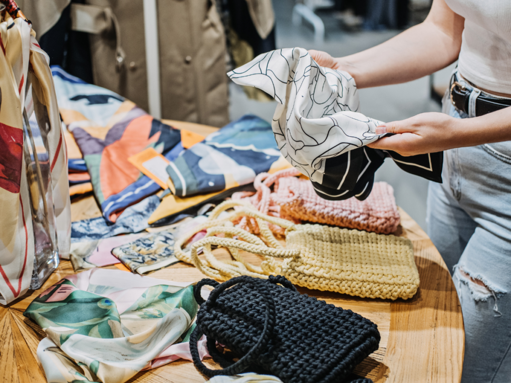 a woman choosing a silk scarf at a store from a table of scarves and purses