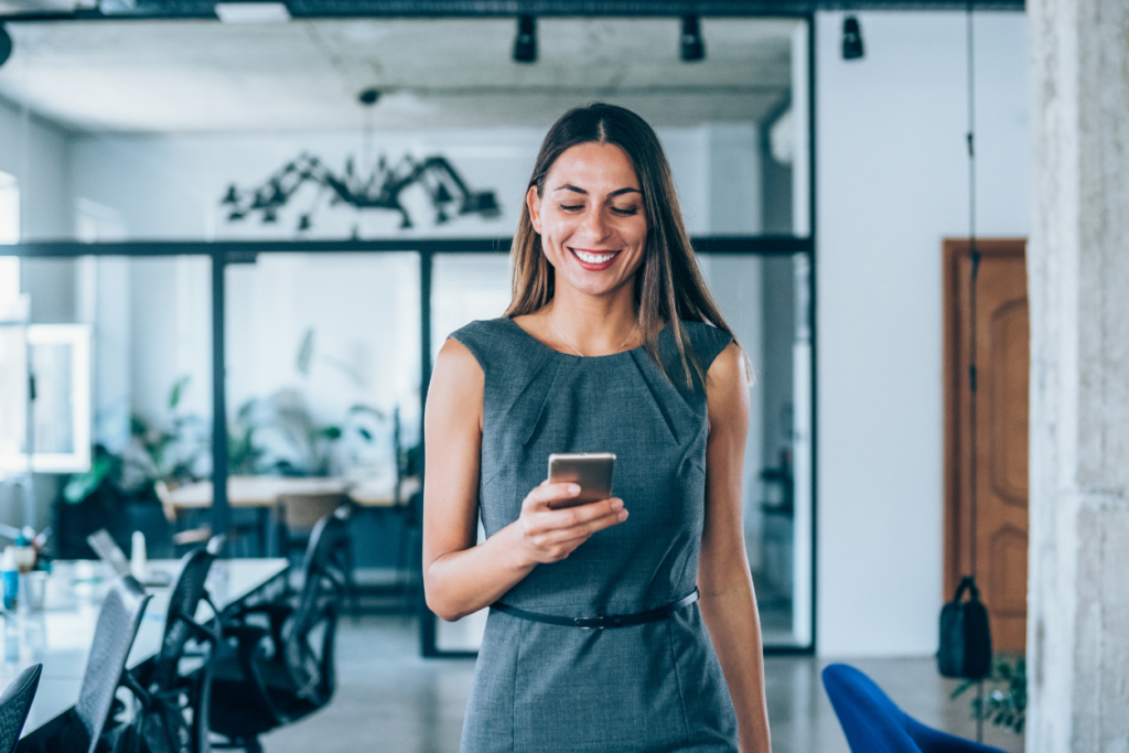 Business woman walking through an office while looking at her phone and smiling